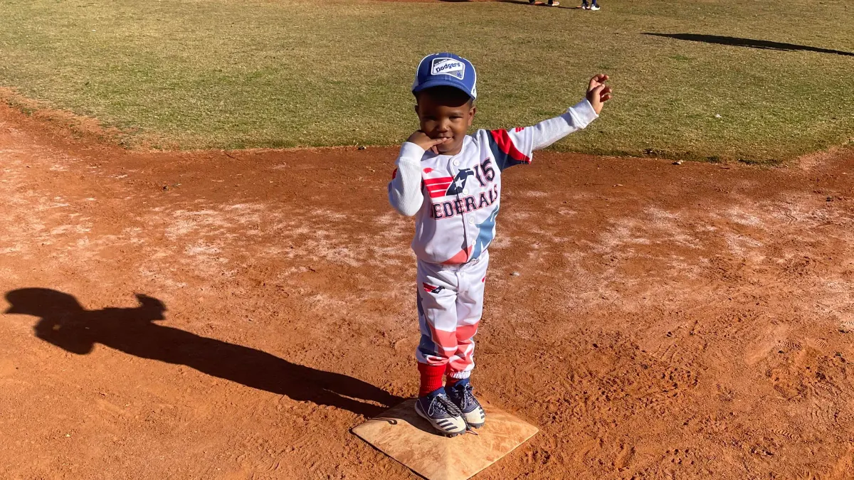 Young baseball player in a Dodgers cap and Federals uniform standing on home plate with one arm raised on a sunny field